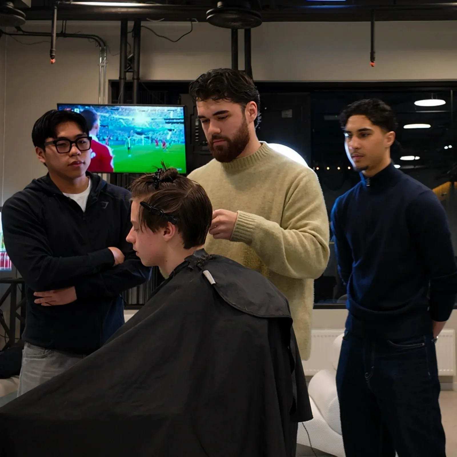 En barber viser klippeteknikk på en kunde mens andre følger nøye med i barberlokalet.